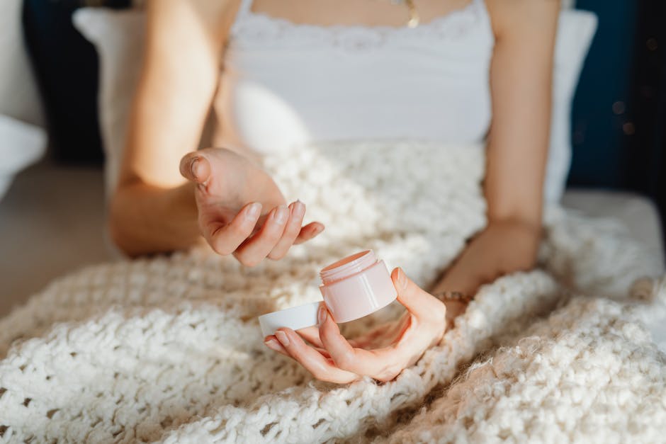 A young woman uses moisturizer while wrapped in a cozy wool blanket indoors.
