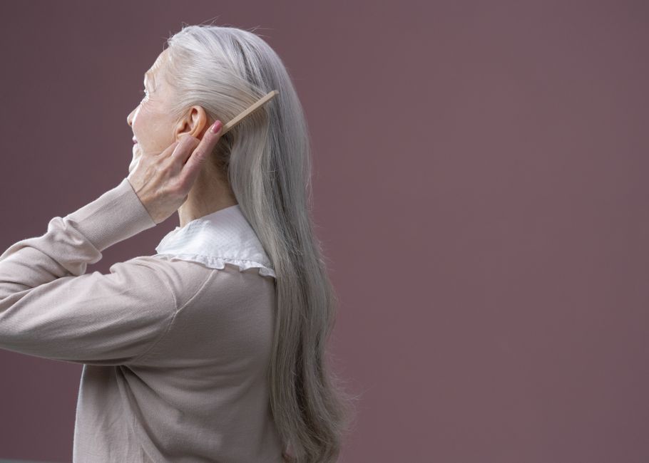 Elderly woman with long grey hair combing it, set against a purple background.