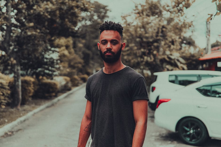 A bearded man with curly hair poses outdoors, wearing a black shirt, showcasing tattoos.