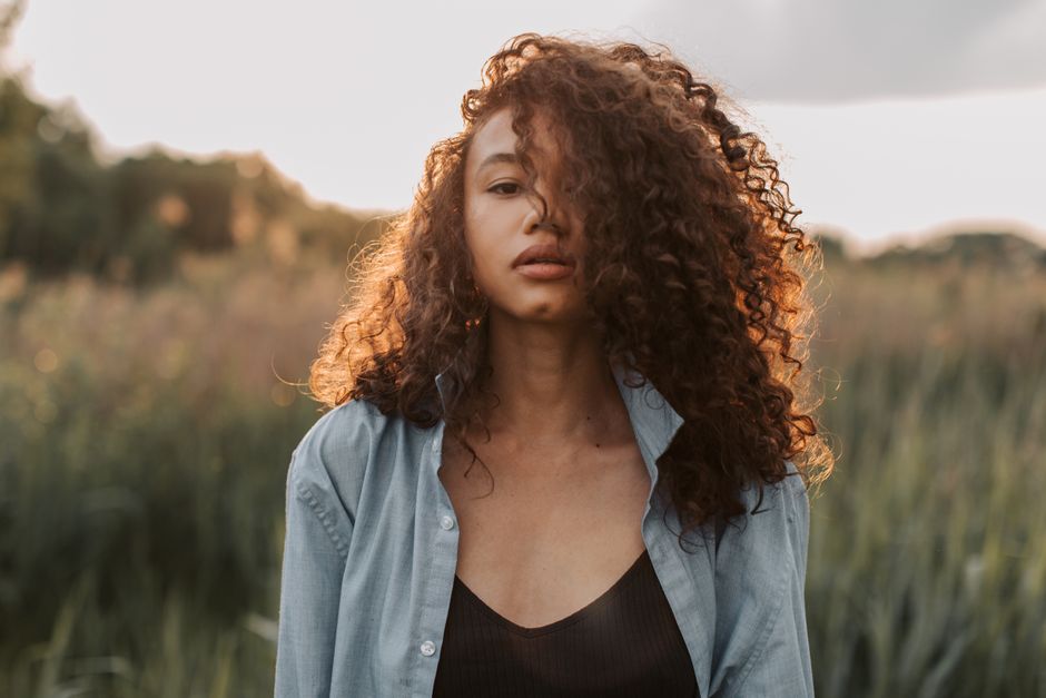 A beautiful woman with curly hair stands stylishly outdoors during sunset.