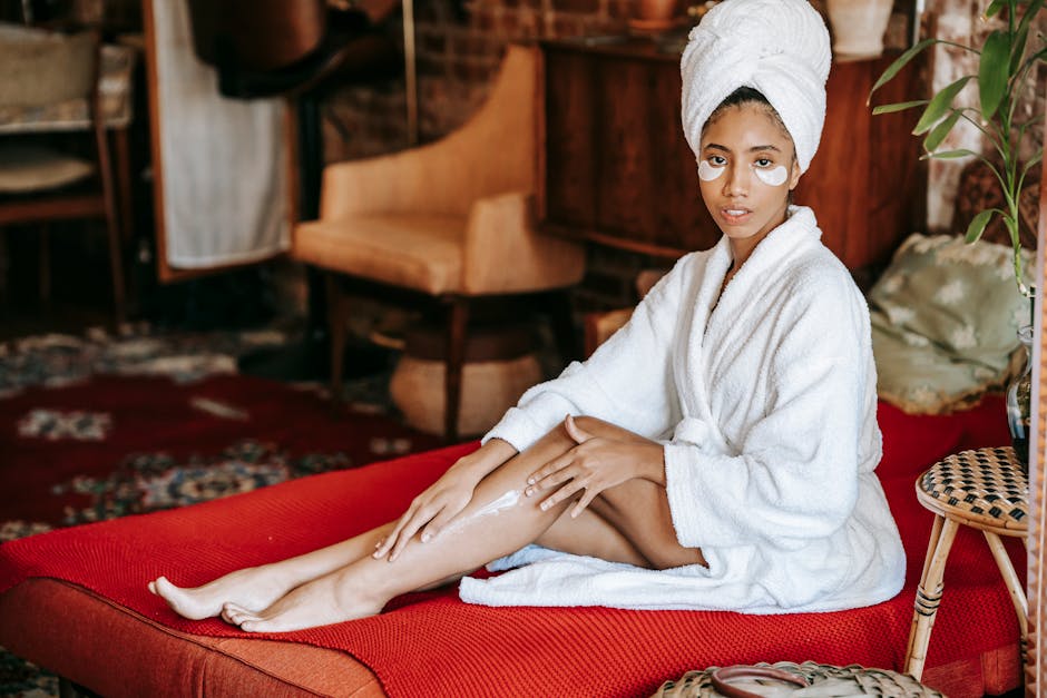 Young barefoot ethnic female in towel and robe applying lotion on leg while sitting on sofa and looking at camera