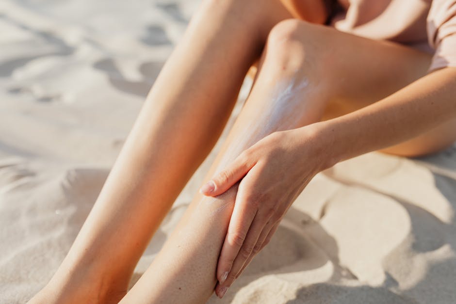 Close-up of a woman applying sunscreen on her legs while sitting on the beach sand.
