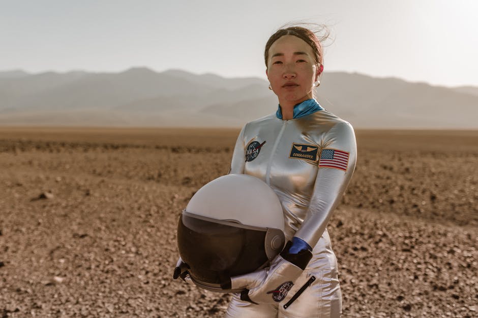 Asian woman astronaut in spacesuit holding helmet in desert terrain.