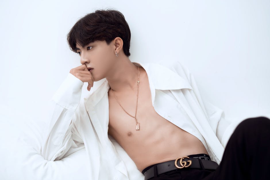 Thoughtful Asian male with dark hair and naked torso in stylish outfit leaning on hand and looking away against white background